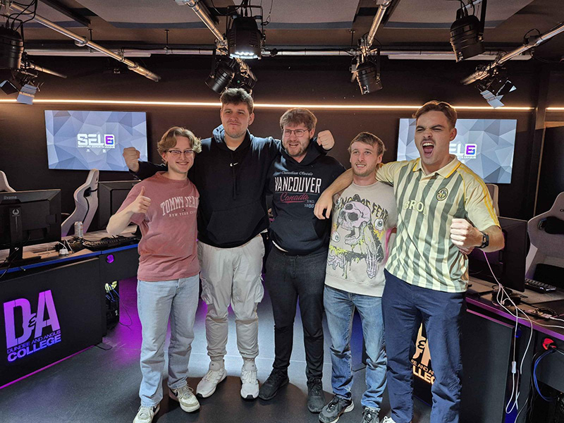 Five individuals celebrating in a gaming room, posing in front of computer stations with raised fists and thumbs up gestures.)